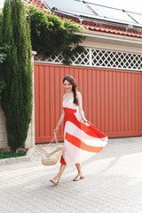 Cheerful young brunette woman, dressed in red and white silk midi striped dress, wicker straw bag, walks along a beautiful green street near the house with a red gate. Outdoor summer fashion portrait.