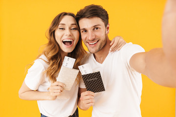 Portrait of happy couple man and woman in basic t-shirts holding travel tickets with passports while taking selfie photo