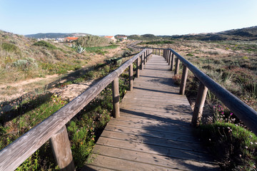 Fototapeta na wymiar Wooden path from sunny beach to village through rural landscape, Portugal. Green hills in peaceful natural area of Algarve