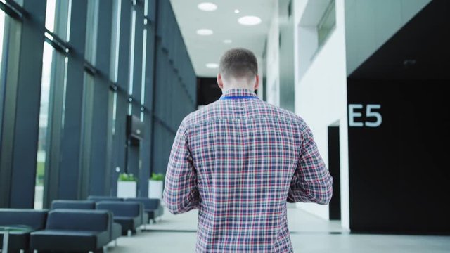 Back view follow shot of young manager with id badge around his neck walking down office building or airport lobby, texting on cellphone, drinking coffee and turning to right, waist up