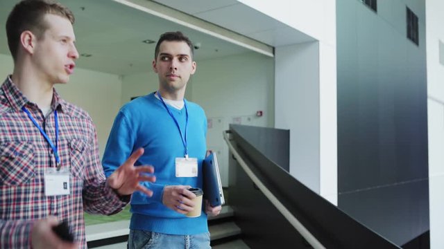 Follow shot of two young business event attendees walking down stairs after conference in modern business center and talking. Men holding laptop computer; takeaway coffee cup and cell phone