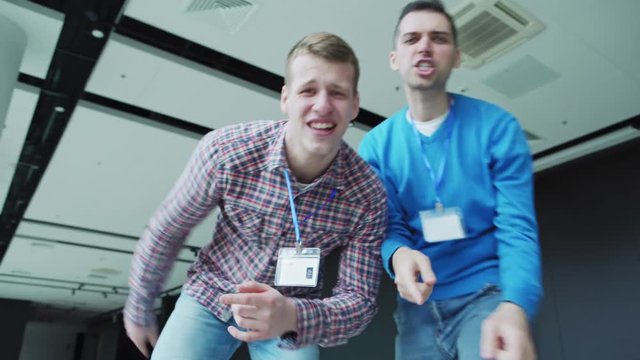 POV low angle handheld shot of two young men with id cards around their necks having fun rapping at camera and dancing in office building. Friends relaxing after business event or shooting music video