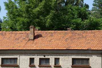 part of the old house with windows under the brown tile roof against the background of branches with green leaves