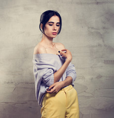 Beautiful young thinking woman posing with hand under the face in violet sweater and yellow pants on grey studio background. Portrait of smiling  natural european girl.