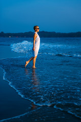 Young and beautiful woman in white dress and sunglasses stands in the surf and looks at blue sea. Figure of girl is reflected in the water.