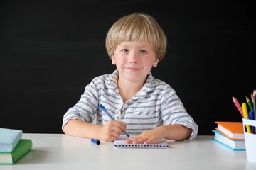 Back to school. Cute little boy sitting at the table on blackboard backgrpund. Child from elementary school. Education concept.