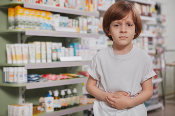 Adorable little boy suffering from abdominal pain, standing alone at drugstore, copy space. Young boy having stomachache