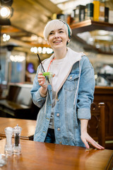 Pretty smiling blonde girl in jeans clothes, holding glass with cocktail in hands. Indoor portrait of joyful young woman in casual clothes laughing in cafe, enjoying cold cocktail in warm day.