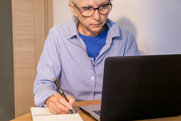 An elderly caucasian woman makes notes in her notebook, looking intently at the laptop screen. Old woman uses modern technology for work