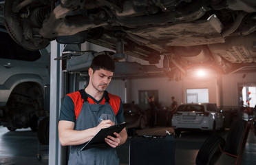 Beautiful lighting behind. Man at the workshop in uniform using notepad for his job for fixing broken car