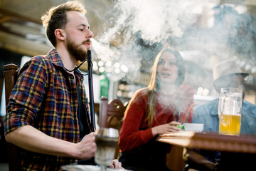 Caucasian young man in casual wear smoking a hookah while sitting at the table in cafe indoors together with friends