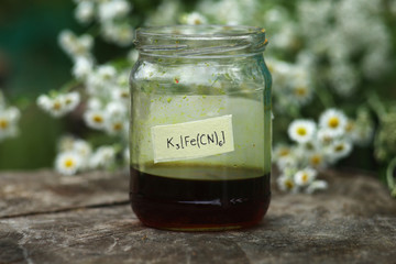 A saturated aqueous solution of the red complex salt of potassium hexacyanoferrate in a glass jar against the background of daisies.