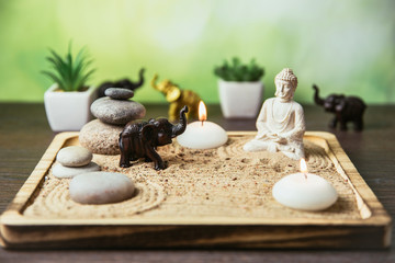 Fototapeta na wymiar Miniature desk zen sandbox with Buddha figure sit in Lotus position, stacked zen sea stones, brown elephant figurines, spa candles burning against green bokeh studio background, copy space.