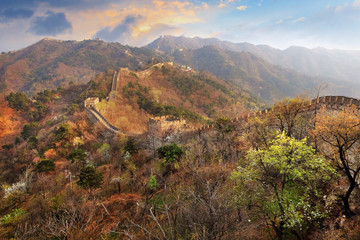 Panoramic view of the Mutianyu section of the Great Wall of China, surrounded by green and yellow vegetation under a colorful sunset light.