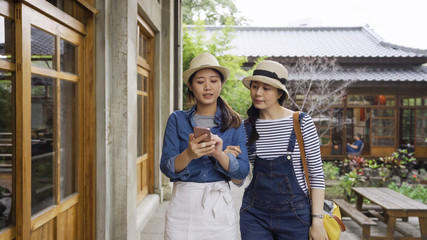 two attractive asian female friends taking walk outdoors smiling looking at cellphone discussing route of tour trip in kyoto japan. girls walking through hallway in garden japanese house in back