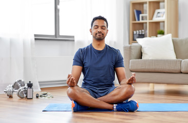 fitness, meditation and healthy lifestyle concept - indian man meditating in lotus pose on exercise mat at home - Powered by Adobe