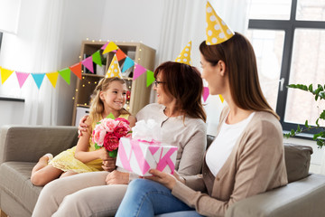 family, generation and celebration concept - smiling granddaughter flowers flowers and mother with gift box greeting grandmother on birthday at home