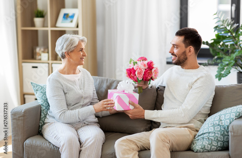 family, mother's day and birthday concept - smiling adult son giving present and flowers to his senior mother at home
