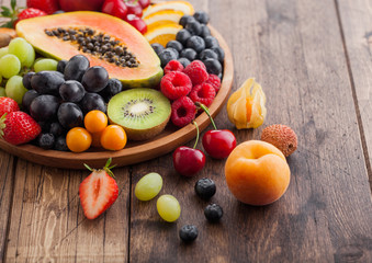 Fototapeta na wymiar Fresh raw organic summer berries and exotic fruits in round wooden plate on wooden kitchen background. Papaya, grapes, nectarine, orange, raspberry, kiwi, strawberry, lychees, cherry. Macro