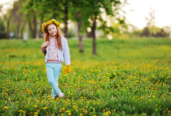 Fototapeta premium a little girl with red hair smiles against a field of dandelions and green grass.