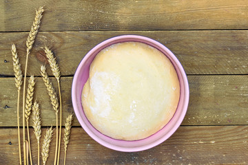 Dough in a pink bowl with wheat spikes on the left on rustic rough wooden background, overhead top view, flat lay, closeup, bread harvesting and pastry concept