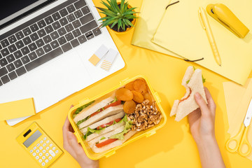 cropped view of woman holding sandwich in hand near lunch box, laptop and office supplies