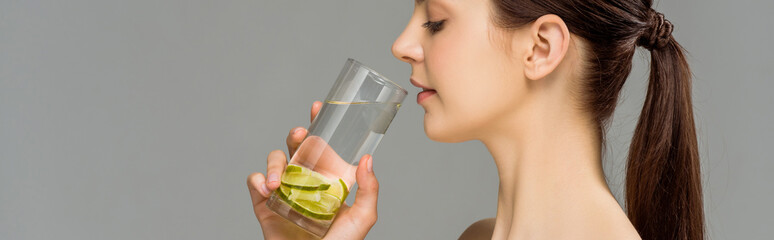 panoramic shot of young woman looking at glass of water with sliced lime isolated on grey