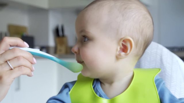 Smiling Baby Boy At Home In High Chair Being Fed Solid Food By Mother With Spoon  