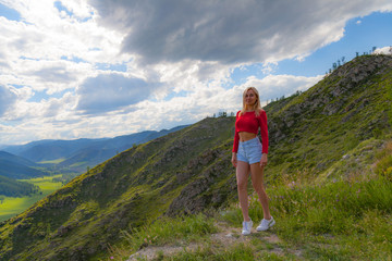 A girl in a red top and blue shorts on the edge of a cliff in the Altai Mountains, below are green fields with trees and grass under a blue sky with clouds.