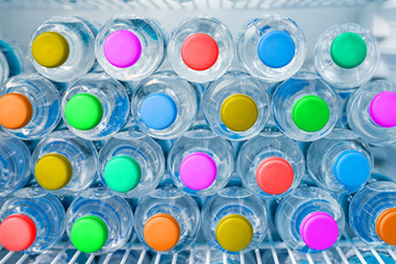 Rows of many transparent plastic drinking water bottles with bright multiclored colorful lids in white refrigerator. Mineral water stack storage in fridge. Healthcare and dehydration prevention