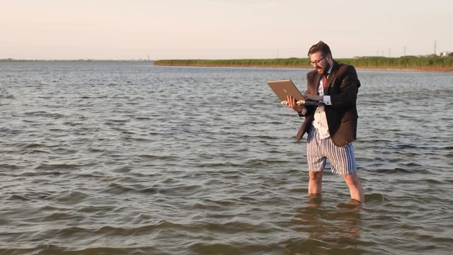 A freelancer with a laptop in a business suit, white shirt and tie, without pants, but in striped men's shorts, in the middle of the sea, is emotionally pleased with the success achieved and raises