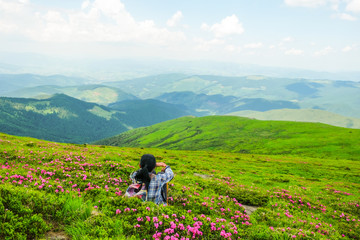 Naklejka na ściany i meble Happy woman 59 years old sits among the flowers of Rhododendron in the Alpine meadows in the Carpathians mountains. Ukrainian tourism. View from the back. Self-isolation concept.