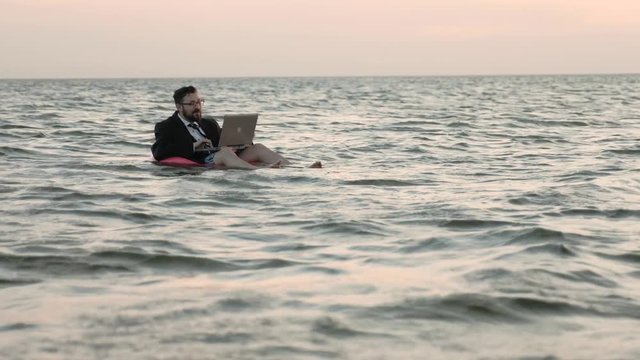 Freelancer floats on the open sea on a pink inflatable circle for children and works with a laptop on his lap.