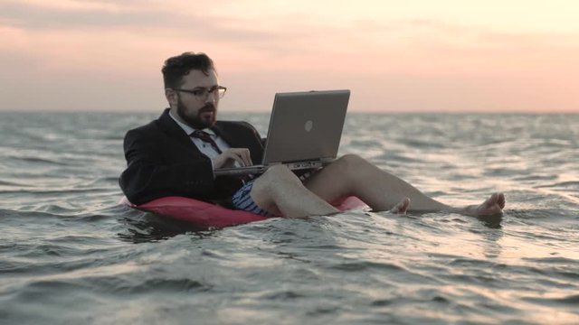 Businessman enthusiastically working behind a laptop swaying on the waves of a reservoir and sitting on a pink inflatable circle.