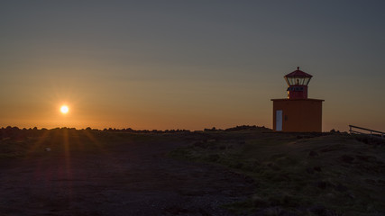 Icelandic  sunset - view of low sun above Atlantic with a lighthouse in foreground. Back light (close to midnight) throws beams on a rocky coastline dominated by a red land orange lighthouse.