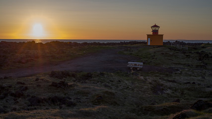 Summer Iceland - sunset into Atlantic with a lighthouse in foreground. Very low sun (close to midnight) throws beams on a rocky coastline dominated by a red lighthouse.