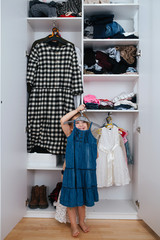 Little jolly girl trying on new dress in front of open wardrobe full of clothes