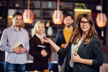 Charming businesswoman in formal wear and with eyeglasses smiling, looking at camera and showing her successful team. Restaurant interior.