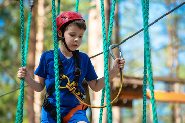 Boy enjoying activity in climbing adventure park at sunny summer day. Kid climbing in rope...