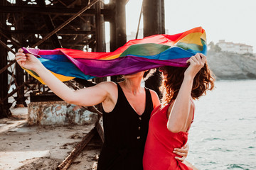 .Young and beautiful lesbian couple proudly celebrating their sexuality at sunset. They hold the rainbow flag of the lgtbi collective, happy and carefree. Love is love.