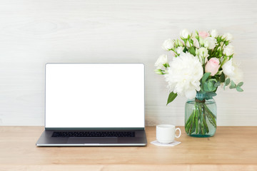 Laptop computer with white blank screen, coffee cup and flowers bouquet on work table. Business and education concept. Feminine home office workspace