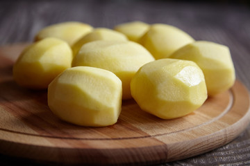 Peeled potatoes on wooden cutting board on brown table. Cooking food from natural products. Root vegetable. Raw ingredient: uncooked whole potatoes