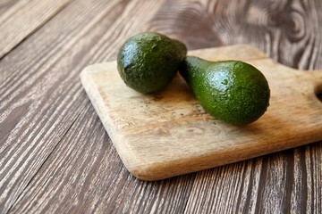 Avocados on a natural pine cutting board on wooden background. Two whole green fresh tropical fruits on brown table