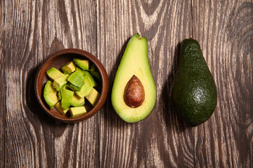 Chopped avocados in a bowl on wooden background. Half avocado with pulp and seed, whole green fresh tropical fruit on brown table, top view