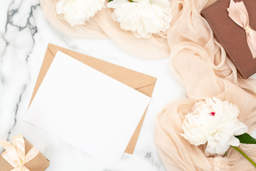 Obraz na płótnie Canvas Top view blank wedding invitation card and craft paper envelope on marble background with white peony flowers and pastel beige scarf. Minimal flatlay style composition, concept of wedding and marriage
