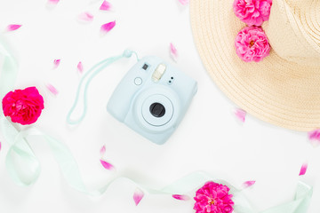 Top view feminine desk with flowers and girl's stuff. Flat lay modern instant camera, staw hat, ribbon, pink rose flowers on white background. Fashion blogger table concept, social media banner.