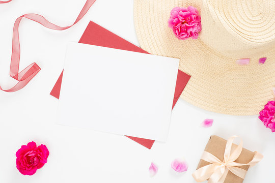 Minimal style composition with blank paper card, craft envelope, beach straw hat, pink rose flowers bud and petal and red ribbon on white background. Flat lay, top view still life concept.
