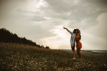 Full length of a amazing couple traveling while man is showing something to her girlfriend against sunset on a field with flowers.