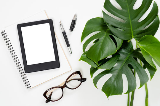 Flat lay tropical jungle Monstera leaves, paper notebook, e-book reader, glasses on white background. Top view feminine diary, stationery and electronic device on home office desk.