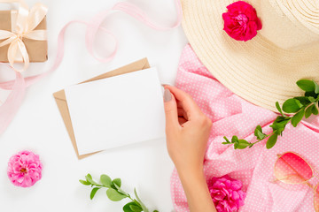 Flat lay fashion office table desk. Female workspace with human hand holding blank paper card, straw hat, rose flower buds, gift box, branches on white background. Top view feminine background